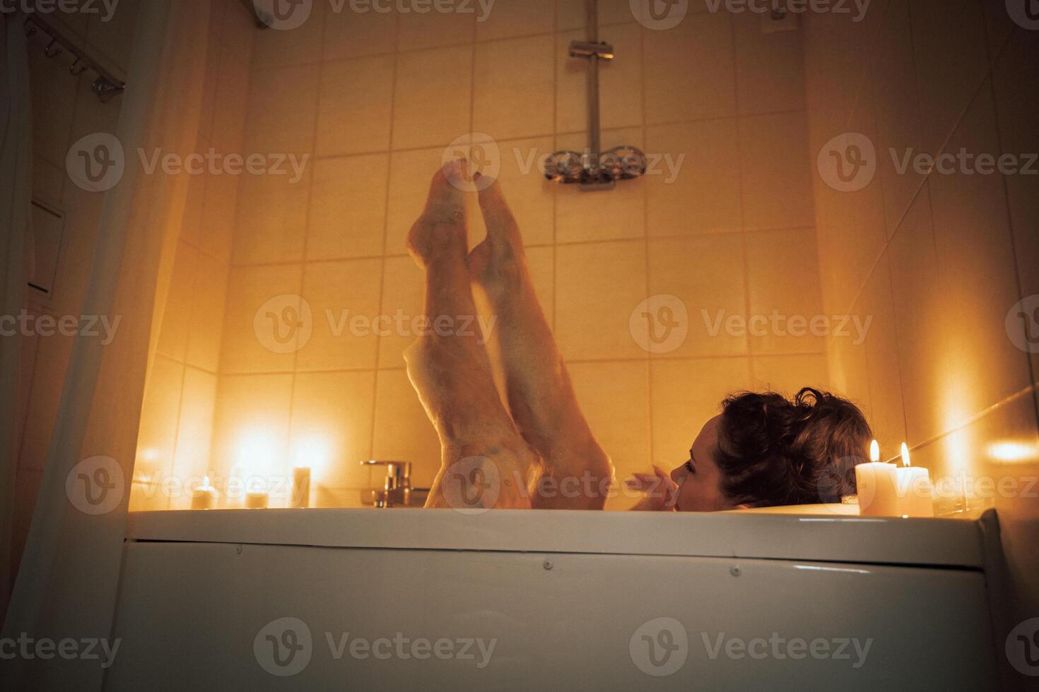 A woman is in a bathtub with candles lit around her. She is barefoot and has her legs up in the air. photo