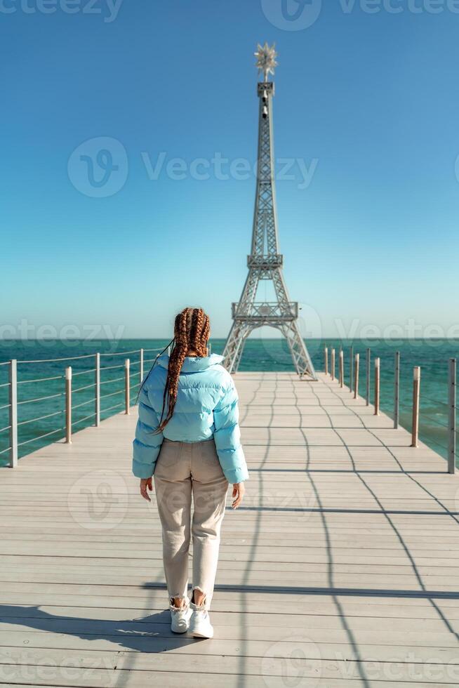 Large model of the Eiffel Tower on the beach. A woman walks along the pier towards the tower, wearing a blue jacket and white jeans. photo