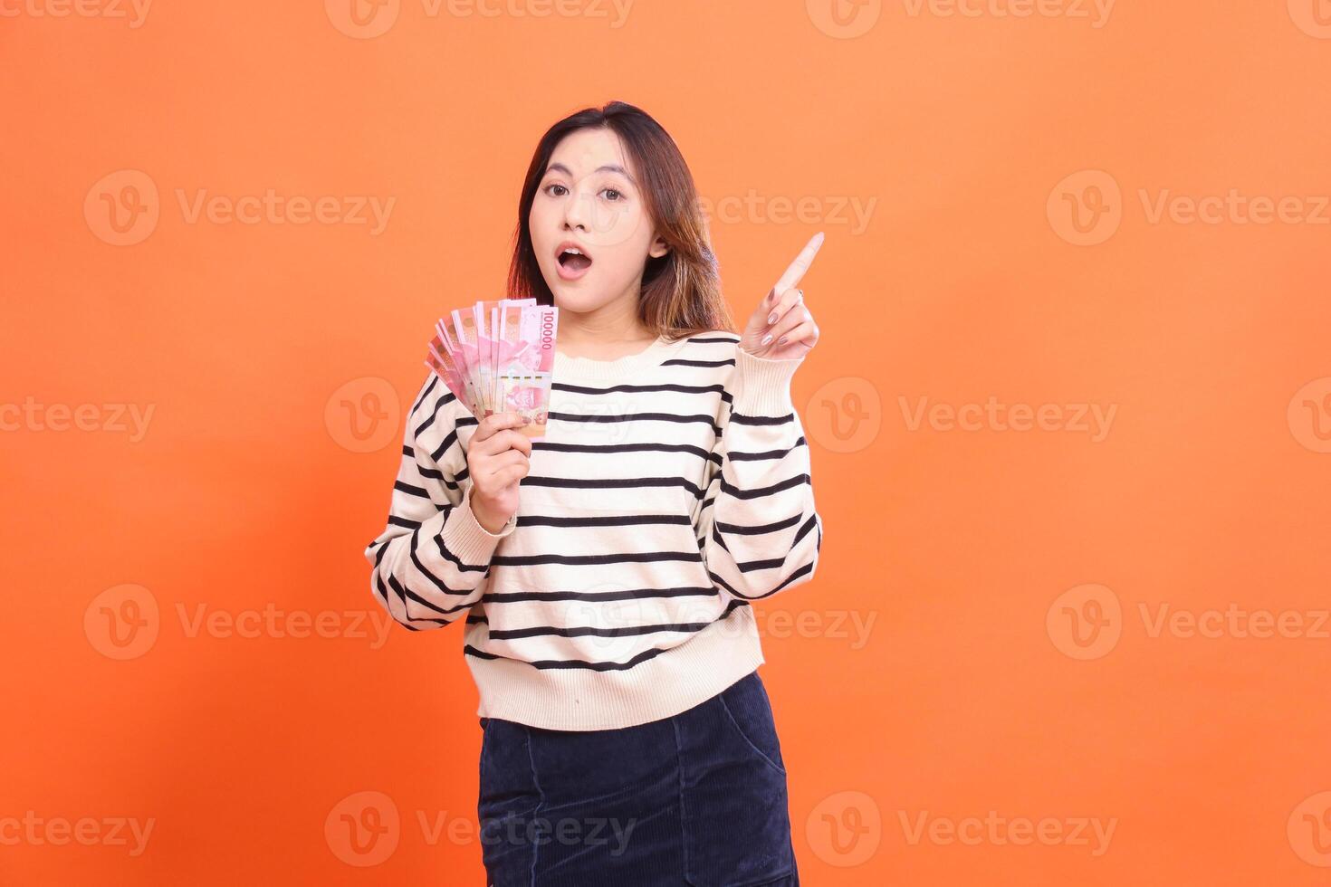 an indonesia woman with a shocked expression at the camera stands holding several rupiah bills while pointing upwards. for fashion, business and finance concept photo