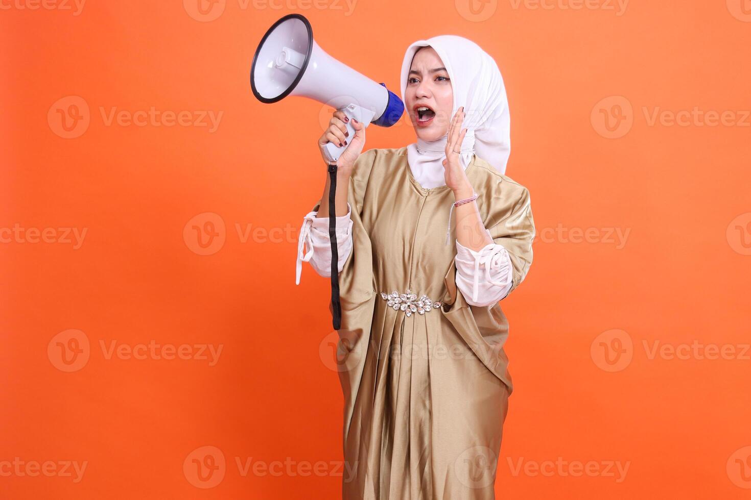 beauty mature indonesia girl shouting candidly holding megaphone loudspeaker palm to mouth wearing kaftan muslimah wearing hijab, isolated on orange background. Fashion concept, broadcast, promotion photo