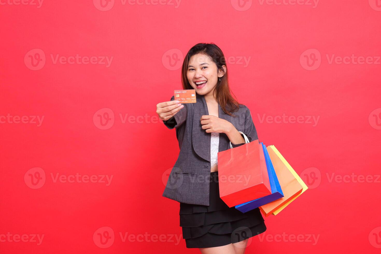 Cheerful Asian office indonesia carrying shopping paper bag while holding credit debit card in front wearing formal jacket with red background. for transaction, lifestyle and business concepts photo