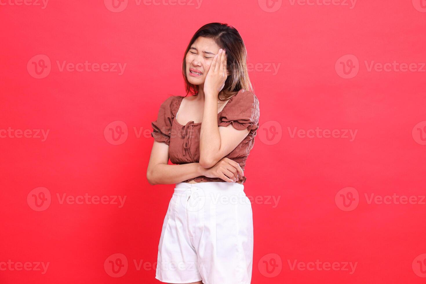 Asian girl expression, holding her head in pain, dizziness, closing her eyes, wearing a brown blouse and shorts with a red background. for health, care and advertising concept photo