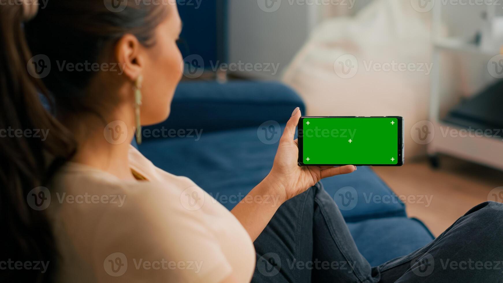 Close up of woman holding smartphone with mock up green screen chroma key display while sitting on sofa in living room. Freelancer using isolated touchscreen device for social networks browsing photo