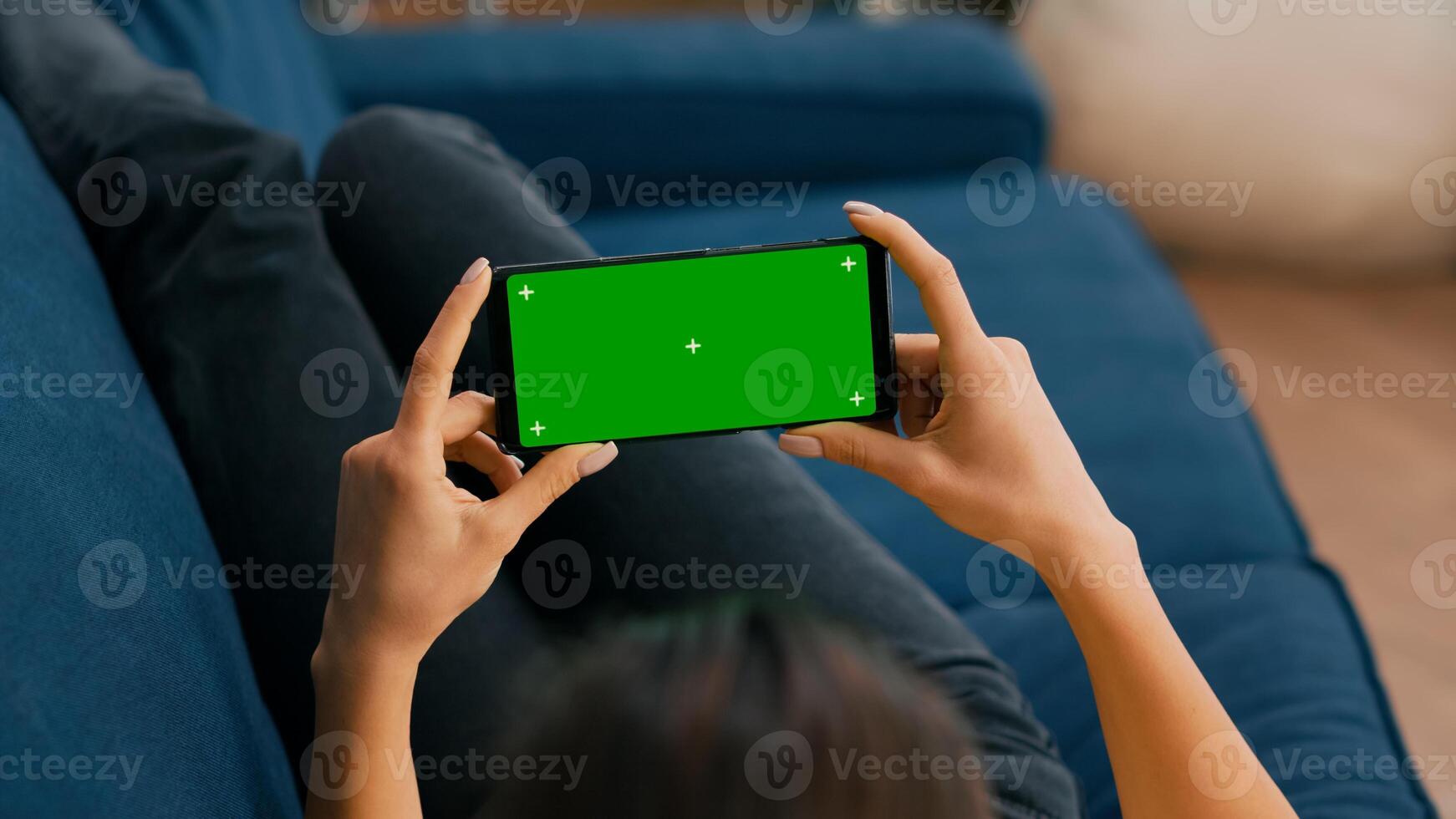 Freelancer sitting on sofa while looking at movies using phone in horizontal mode with mock up green screen chroma key display. Woman using isolated touchscreen device for social networks browsing photo