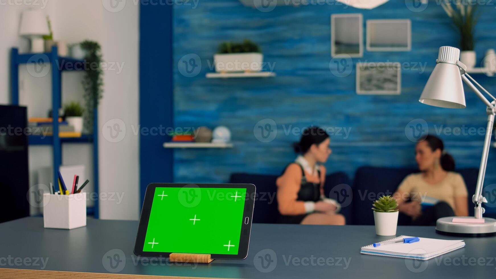 Tablet computer with mock up green screen chroma key display standing in front on office desk. Two collegues talking in background, sitting on sofa about internet connection photo