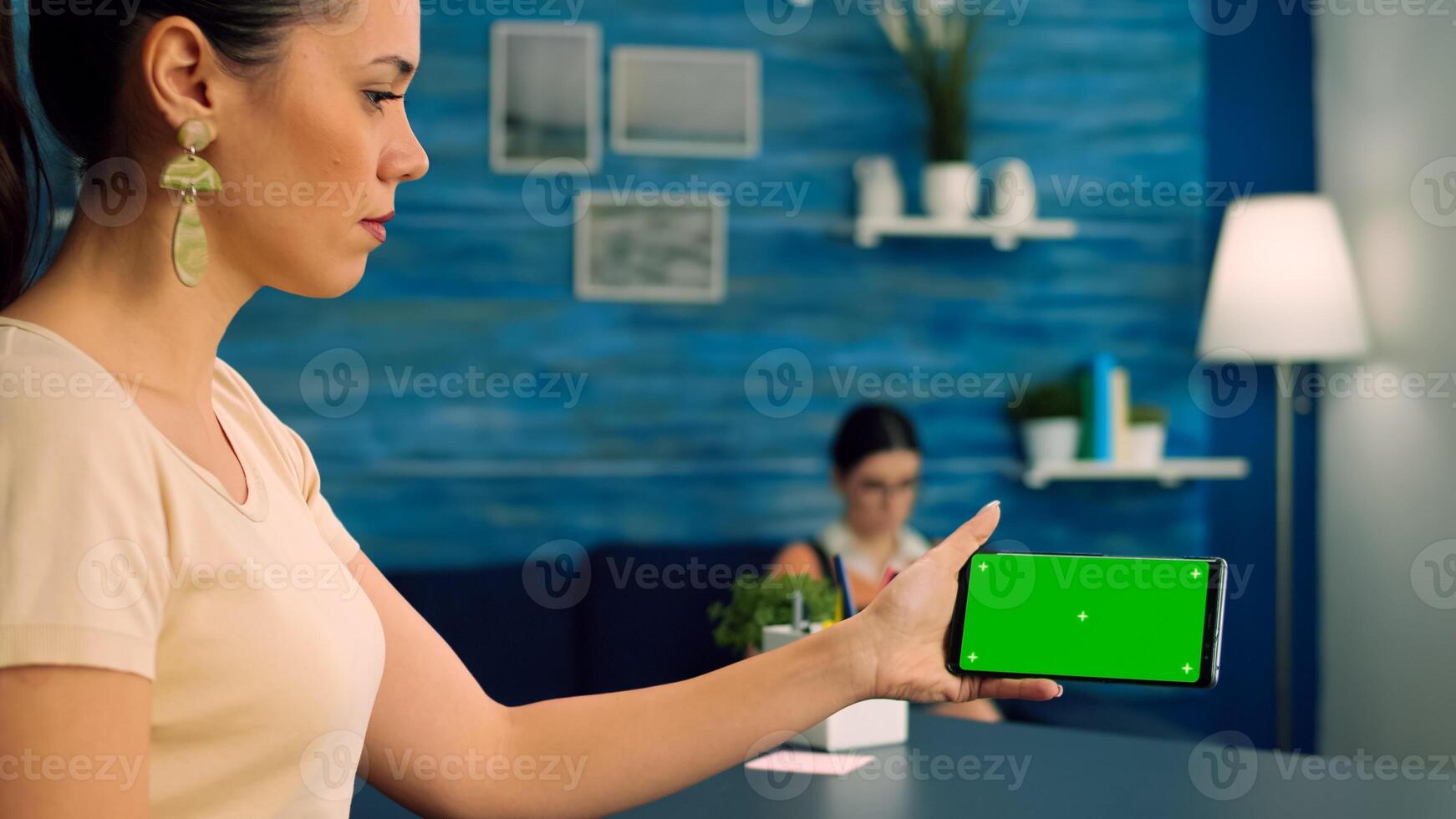 Caucasian female holding isolated smartphone with mock up green screen chroma key display, browsing online information on internet while her collegue reading a book in background photo