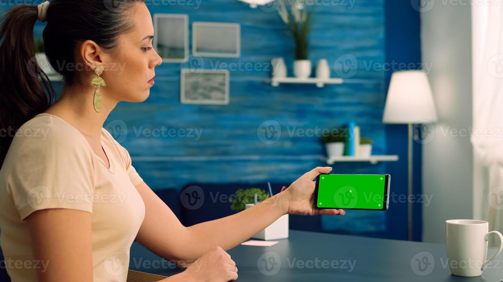 Social media person holding phone with mock up green screen chroma key display while watching online videos. Caucasian female sitting at office desk in living room using isolated technology device photo