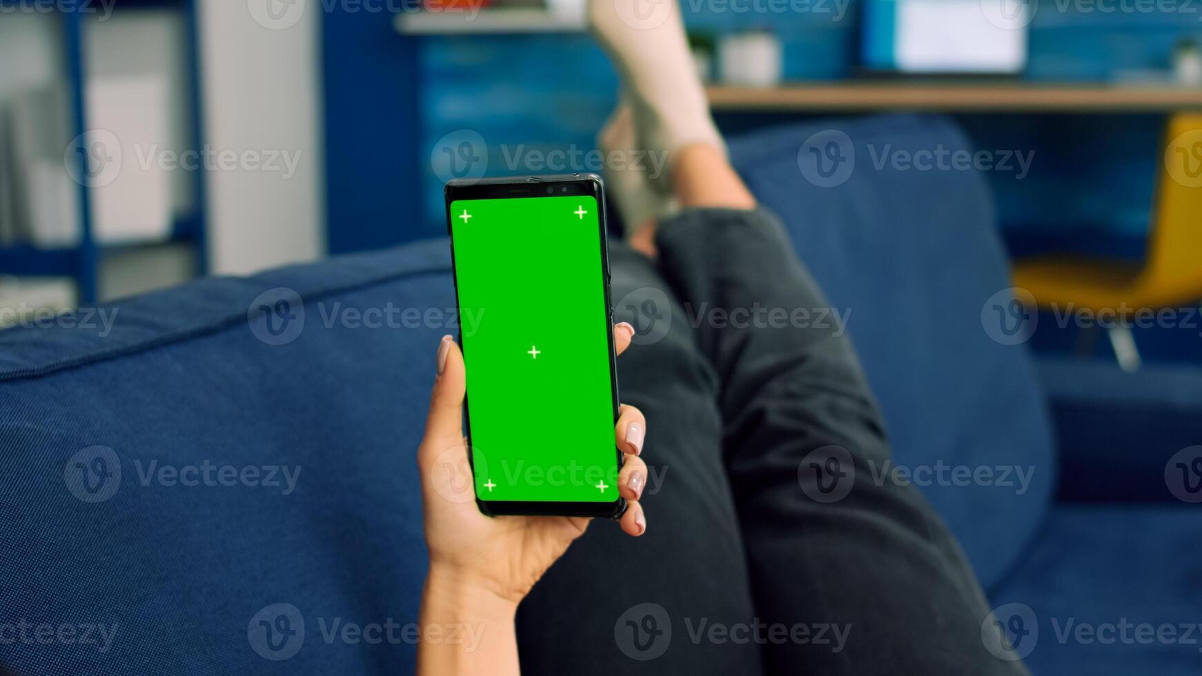 Freelancer woman lying on sofa looking at mock up green screen chroma key smartphone in vertical mode. Caucasian female browsing on internet using isolated phone for social media network photo