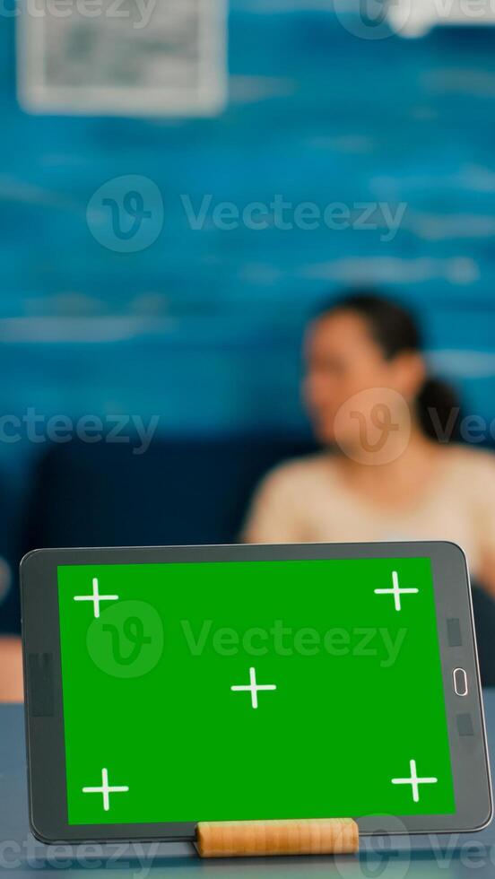 Isolated digital tablet with mock up green screen chroma key display standing on office desk in living room. In background room collegues talking about online communication photo