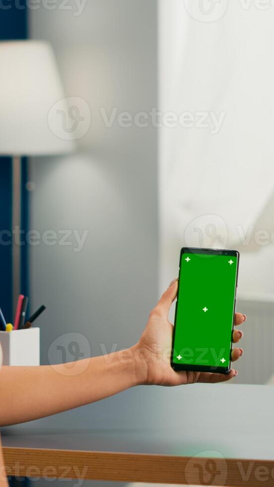 Social media woman holding touchscreen phone with mock up green screen chroma key sitting at modern office desk. Caucasian female working at online app using smartphone with isolated display photo