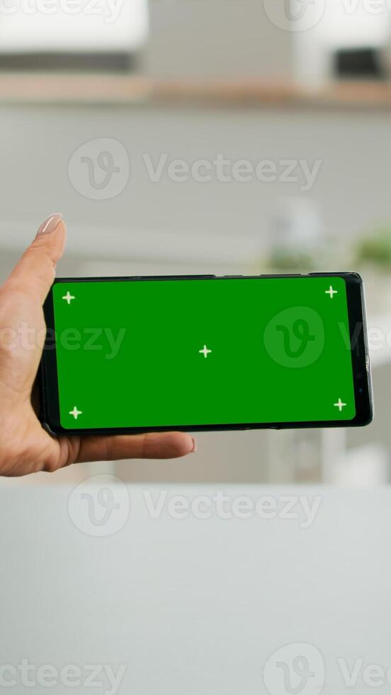 Freelancer holding smartphone with mock up green screen chroma key in horizontal position. Business woman searching online information using isolated device sitting on office desk photo
