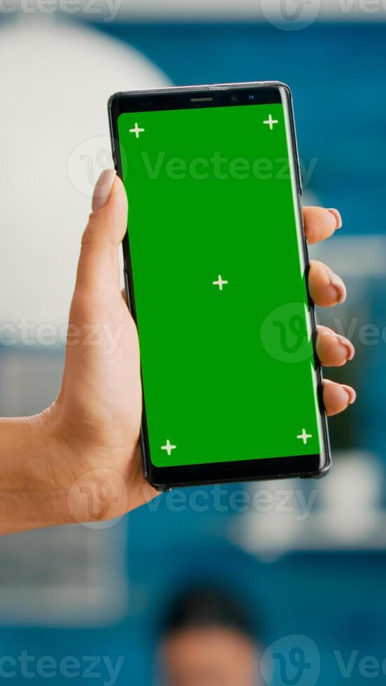 Hands of freelencer holding vertical mock up green screen chroma key smartphone. Business woman using isolated phone for browsing social networks sitting on office desk photo