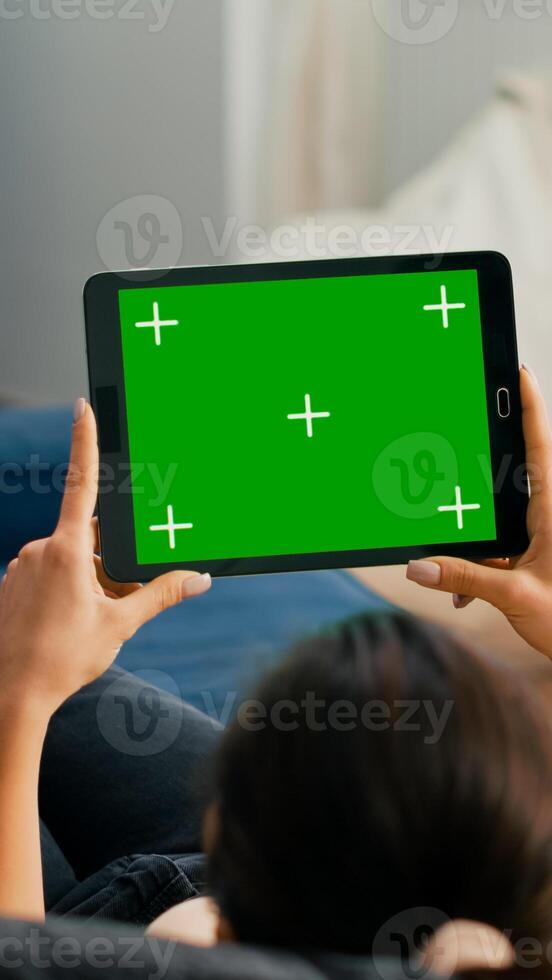 Woman sitting on sofa in living room watching movies on tablet computer with mock up green screen chroma key display. Freelancer using isolated touchscreen device for social networks browsing photo
