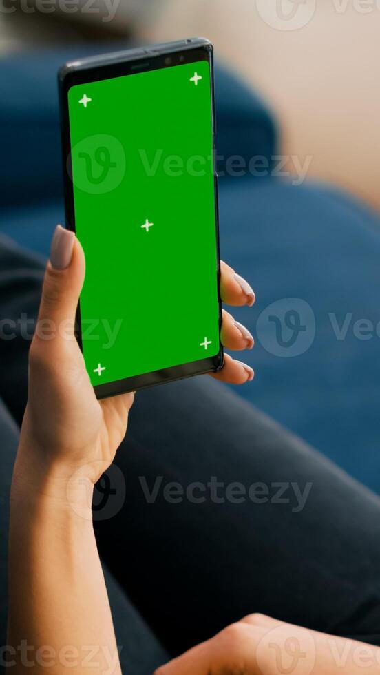 Freelancer holding in vertical mode smartphone with mock up green screen chroma key display while lying on sofa in living room. Woman using isolated touchscreen device for social networks browsing photo
