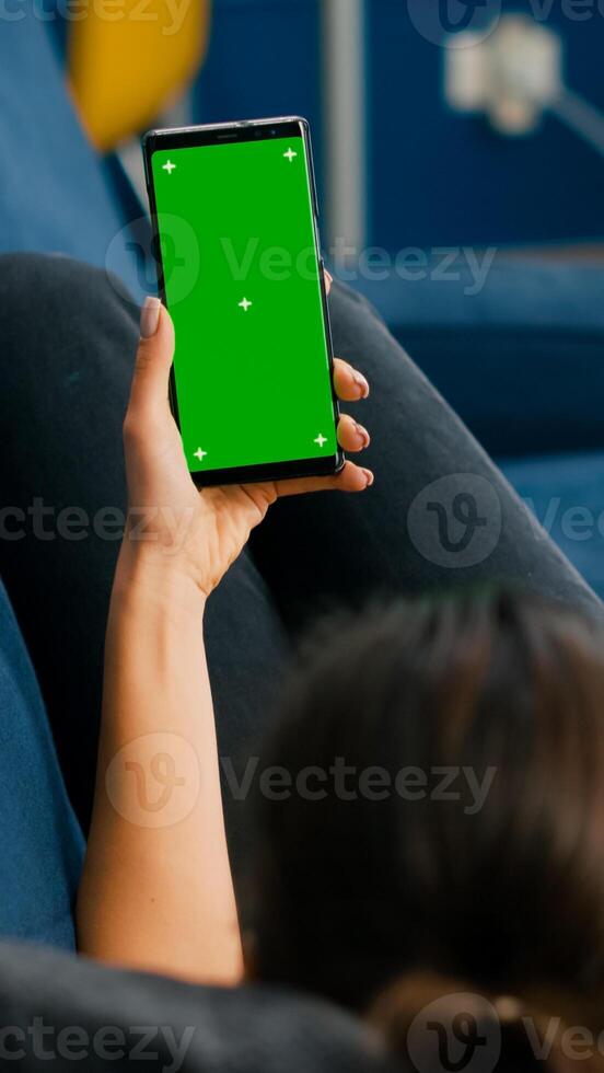 Freelancer lying on sofa while having online videocall on smartphone with mock up green screen chroma key display in vertical mode. Woman using isolated touchscreen device for social networks browsing photo