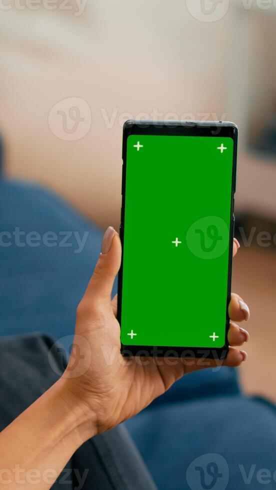 Caucasian female sitting on sofa in living room while socializes on internet app using phone with mock up green screen chroma key display.Woman browsing on network with isolated gadget photo