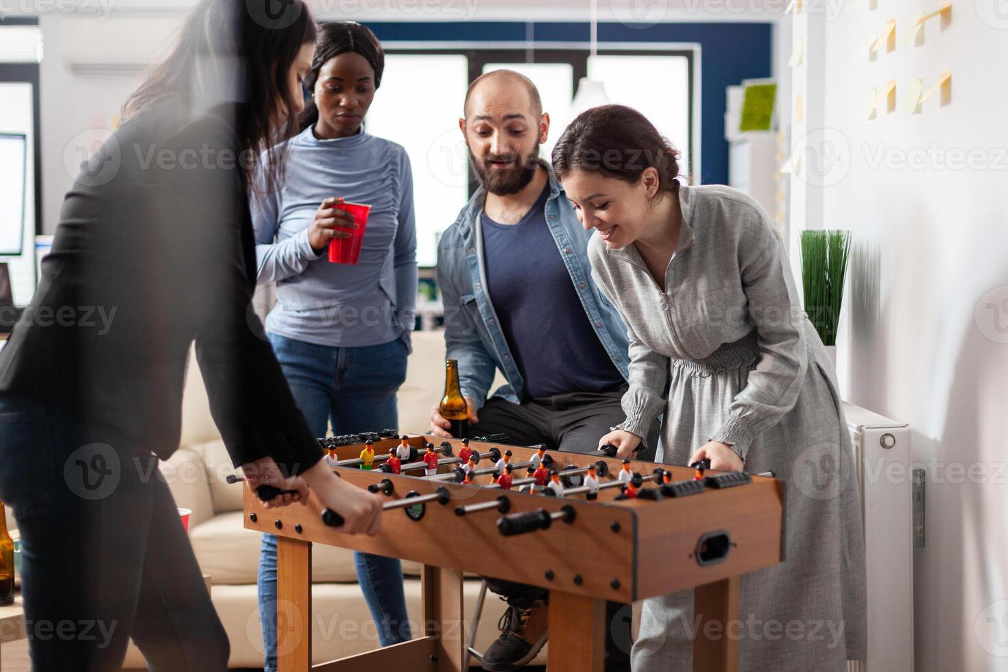 Multi ethnic group of people playing at foosball table after work partying and enjoying alcohol cups bottles of beer. Diverse team colleagues celebrating break from business activities photo