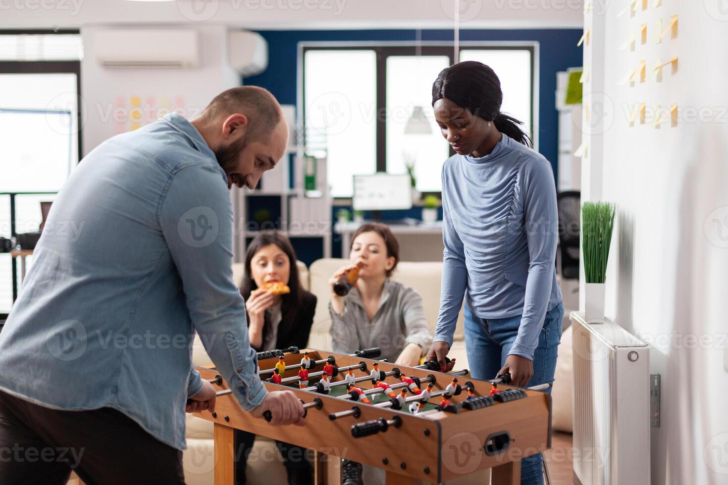 Multi ethnic friends enjoy after work foosball game at table soccer football while holding cups bottles of beer alcohol. Diverse group of colleagues having fun entertainment enjoyment photo