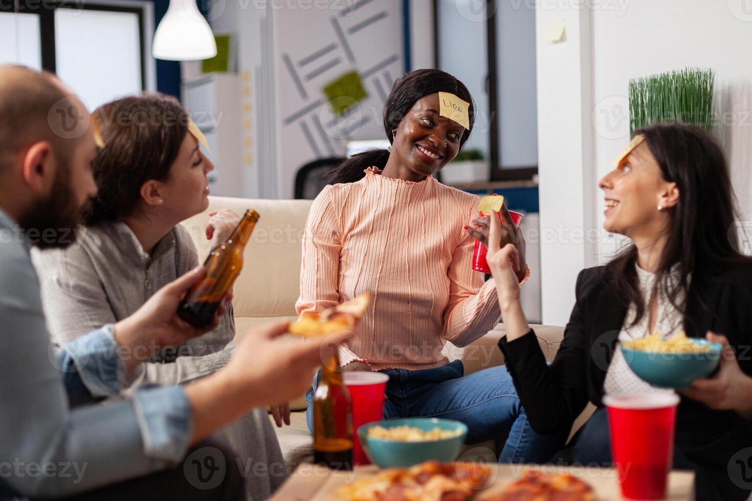 African american woman playing guessing game with friends after work at office. Multi ethnic group of workers guess imitation for fun cheerful activity while eating and drinking beer photo