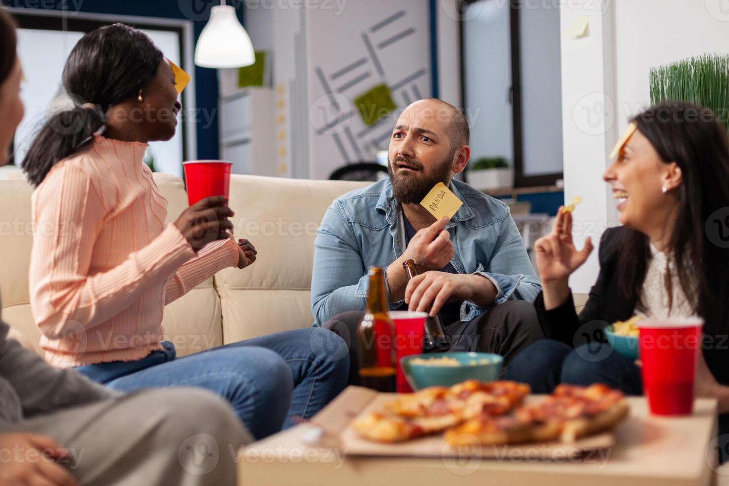 DIverse team of coworkers playing charades after work at office space. Multi ethnic friends enjoy guessing game of imiation as fun cheerful activity entertainment with food and drinks photo