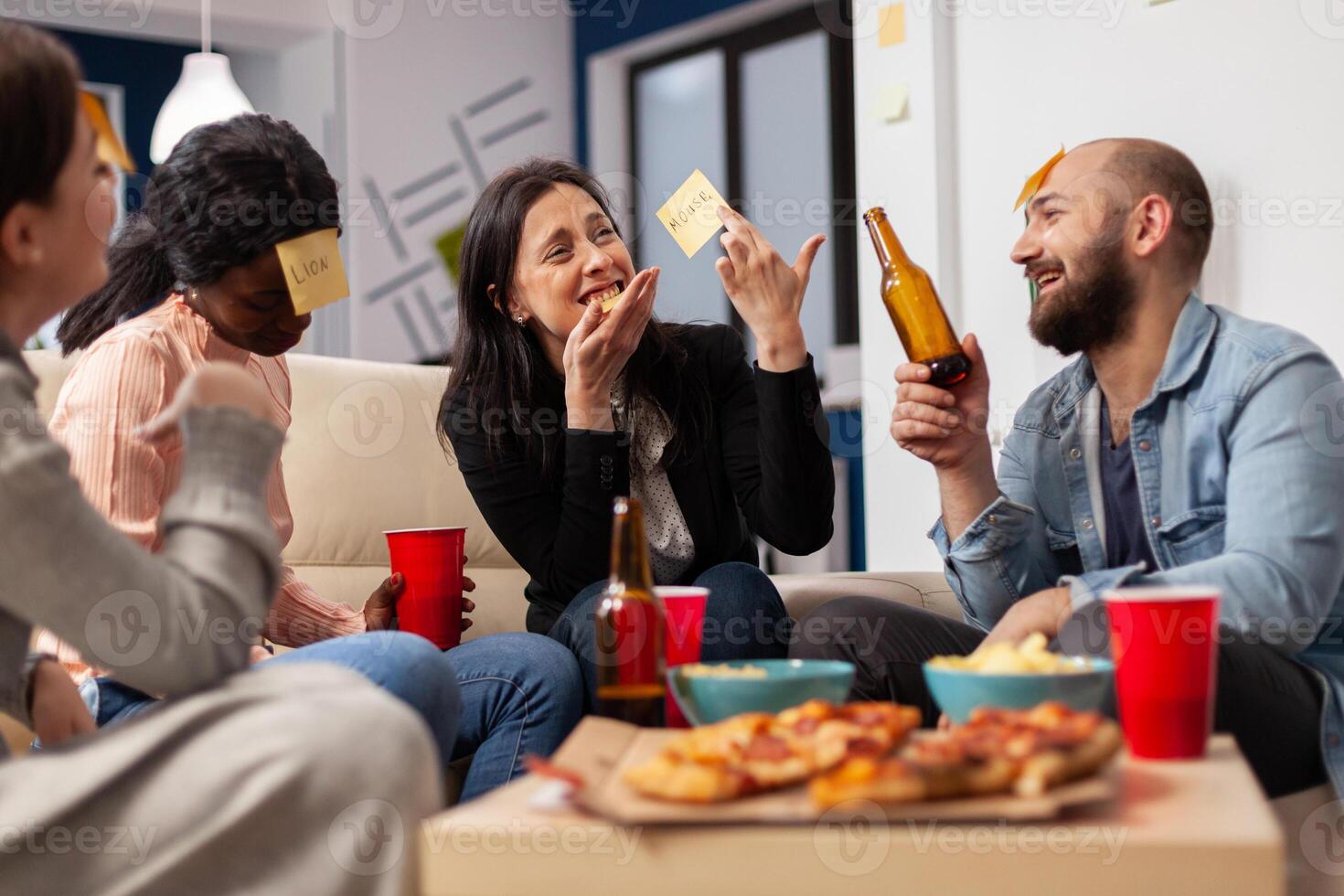 Cheerful group of colleagues enjoying a game of charades after work at office. Multi ethnic people play imitation concept for fun activity entertainment while eating pizza and drinking beer photo