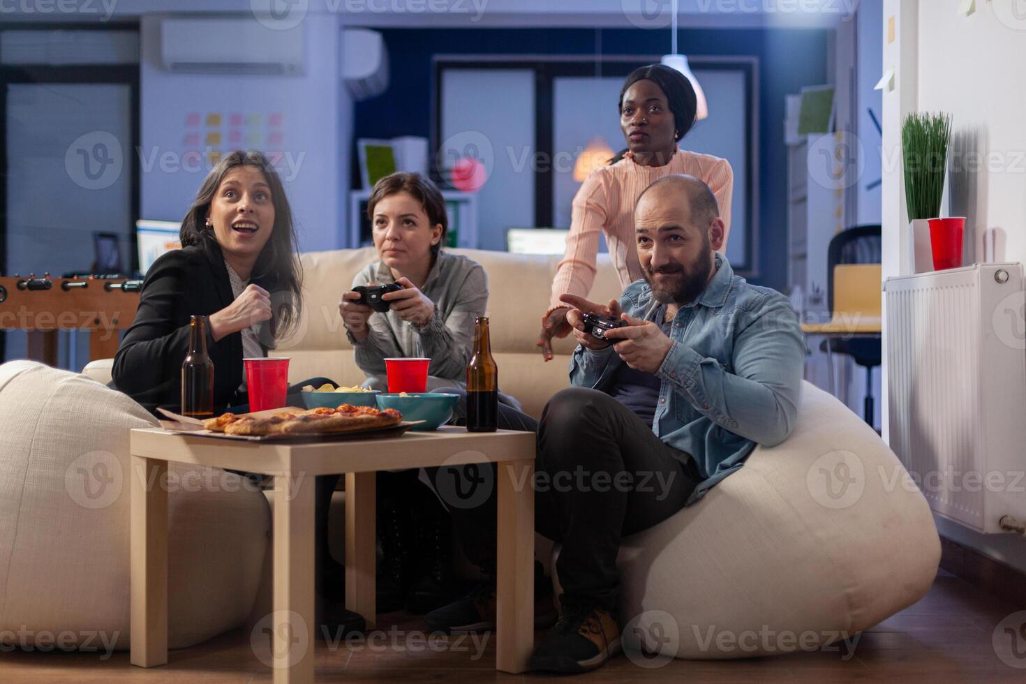 Multi ethnic group of coworkers play game on console while holding joystick. Cheerful diverse friends enjoy fun competition on tv computer while having snacks and beer of table photo