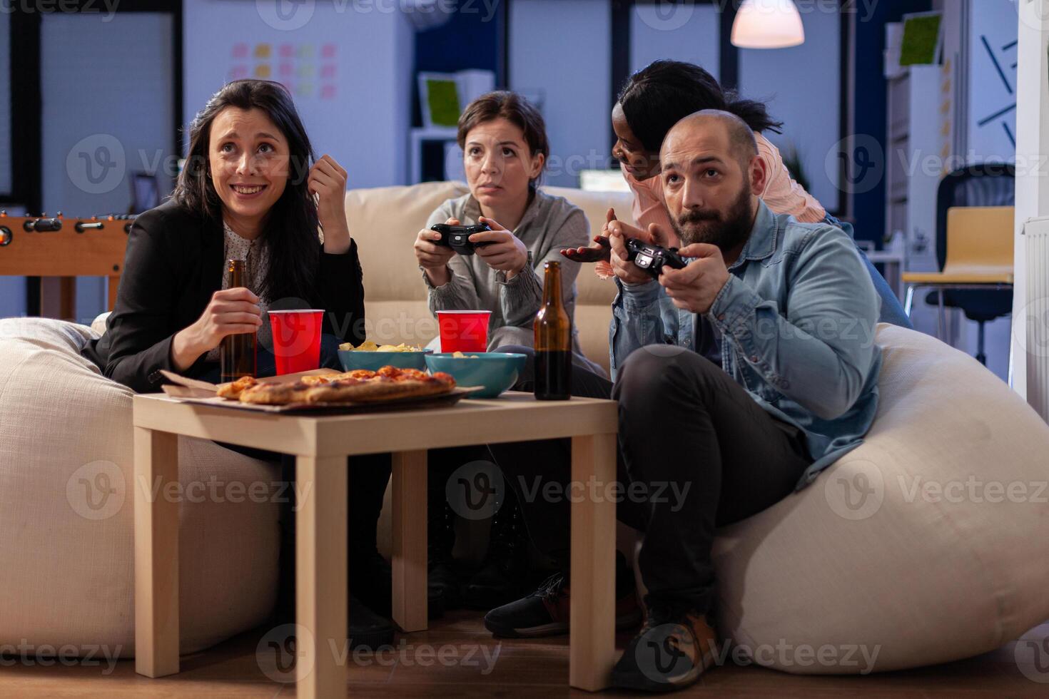Diverse team of workers playing console game on tv at office after work using joysticks on sofa. Multi ethnic friends starting competition while having snacks pizza chips and drinking beer photo