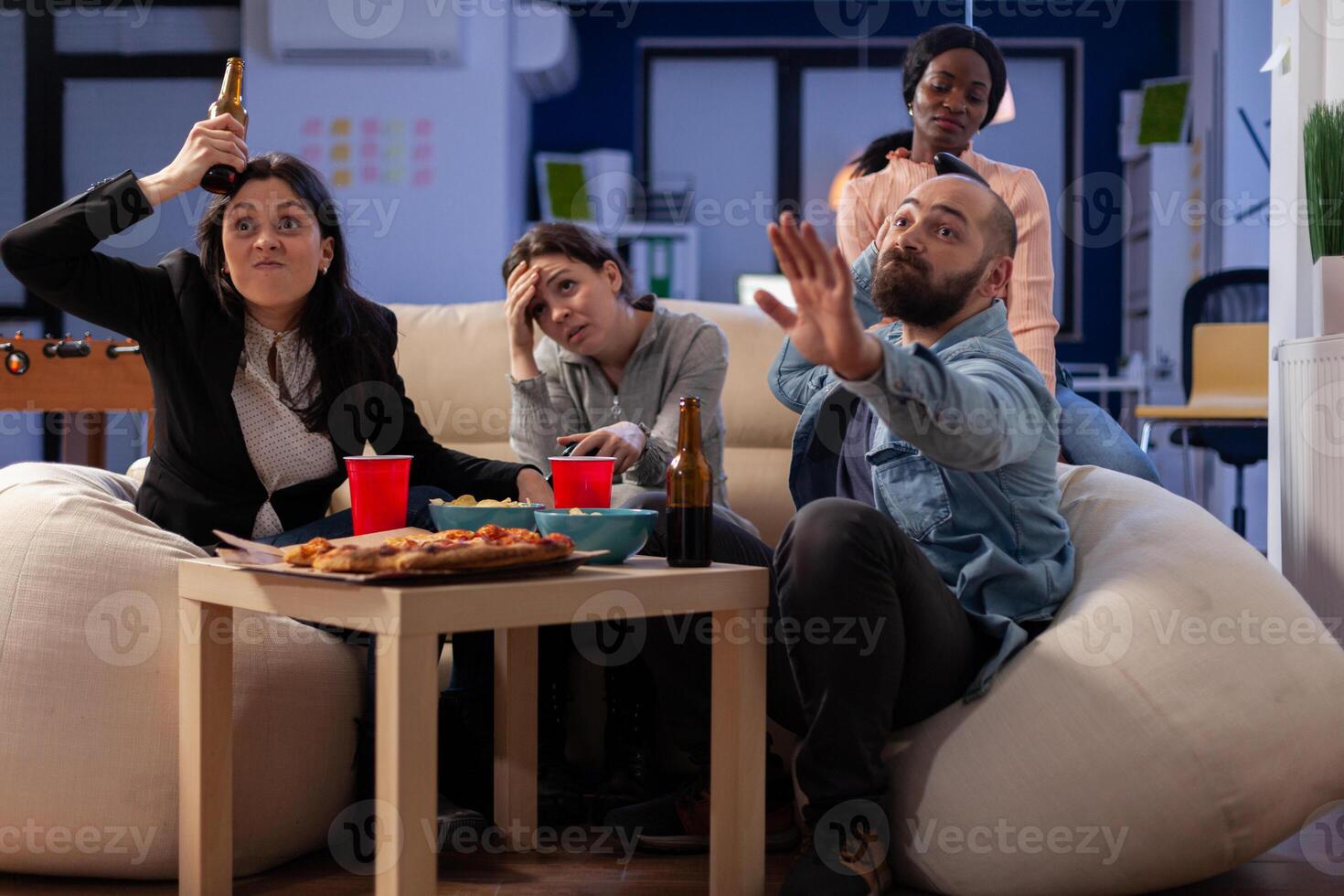 Team of multi ethnic friends losing tv game on console after work at office. Cheerful diverse group play with joystick while eating snacks pizza chips and drinking beer alcohol from cups photo