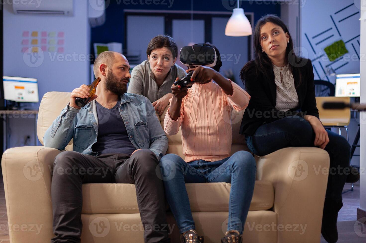 Group of multi ethnic friends using vr glasses for fun after work at office while holding controller joystick. Diverse team enjoy celebration party bonding together with drinks and snacks photo
