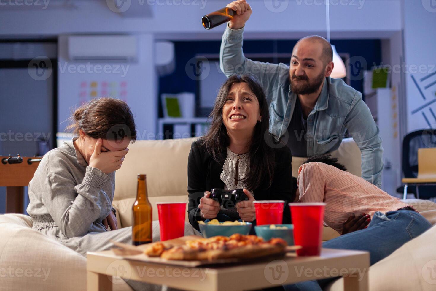 Diverse group of mates bonding while playing game on tv console losing with joystick controller after work. Multi ethnic team enjoy office celebration party with snacks and drinks photo