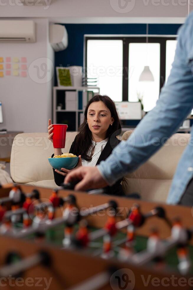 Portrait of young woman holding glass of beer at after work celebration, looking at foosball game play. Cheerful person enjoying drinks part after hours, celebrating with office workmates. photo