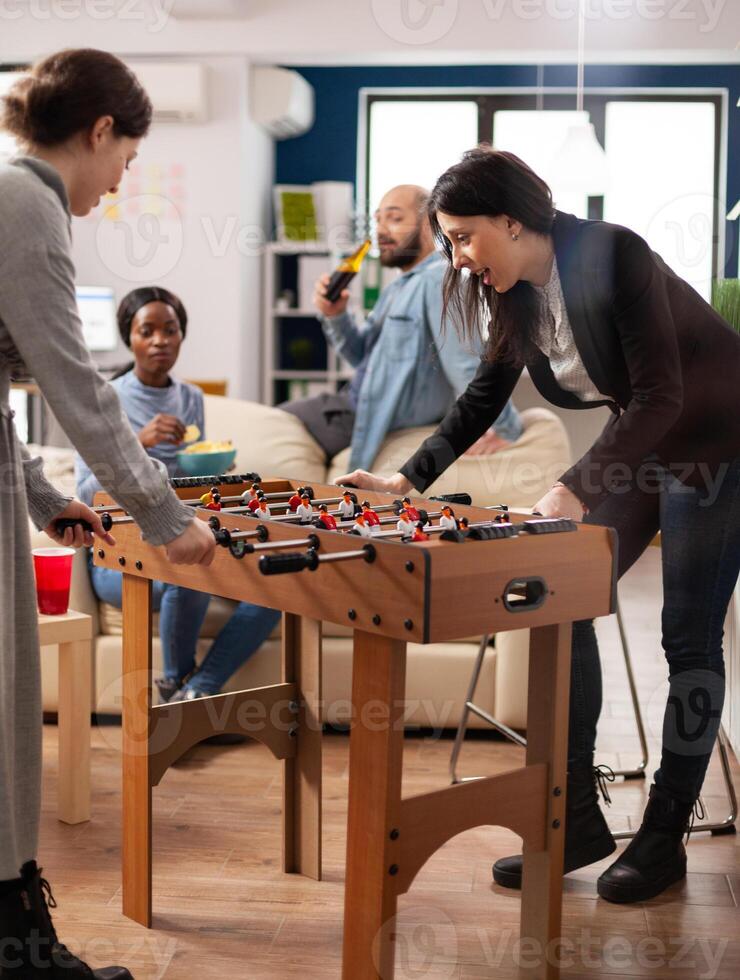 Multiethnic group of people playing at foosball game table, enjoying soccer match with toy in business office. Coworkers having fun with after work alcoholic drinks, snacks and competition. photo