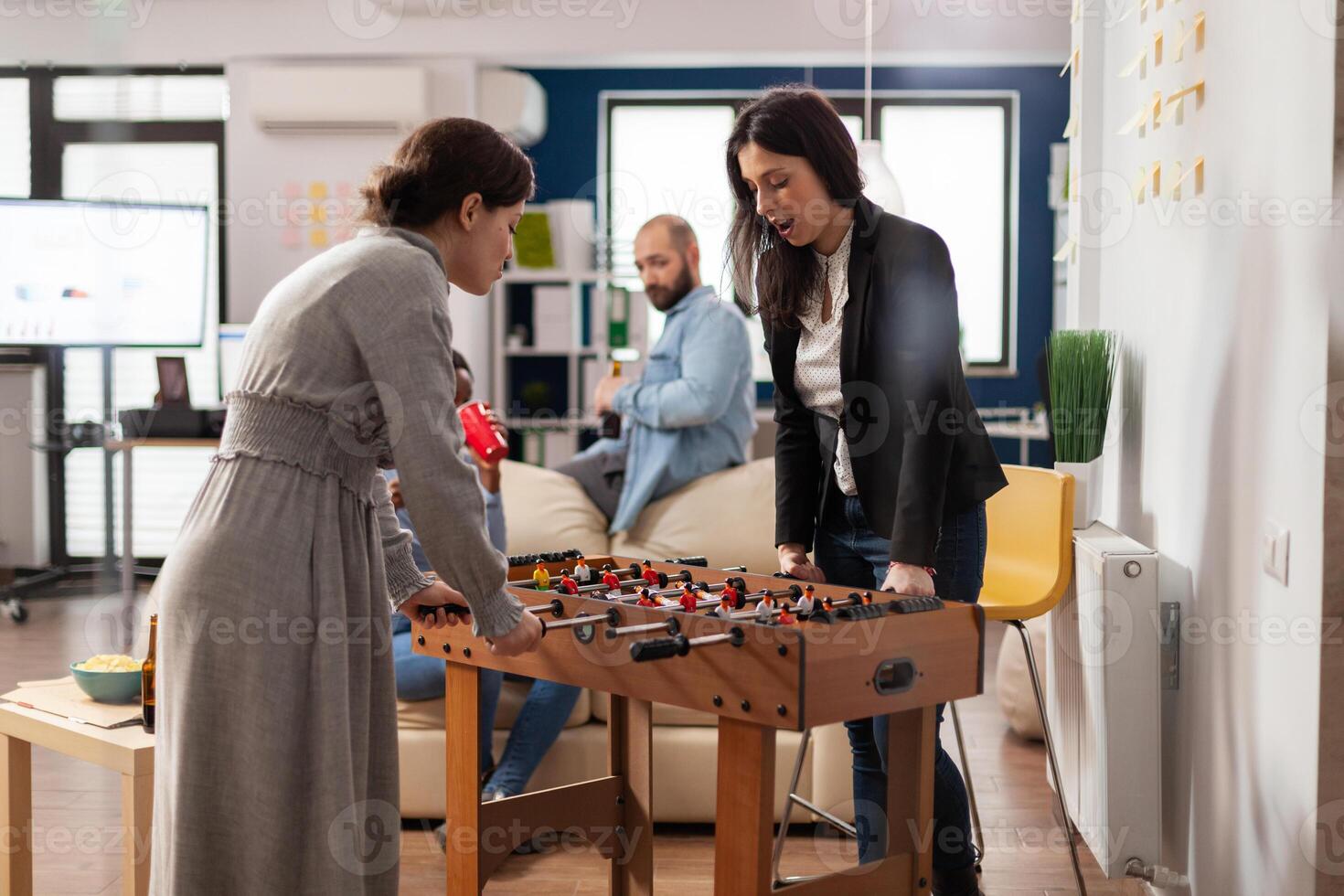 Diverse team of coworkers playing foosball game table match, celebrating with drinks after work hours. Workmates enjoying party celebration with beer, pizza and soccer in office. photo