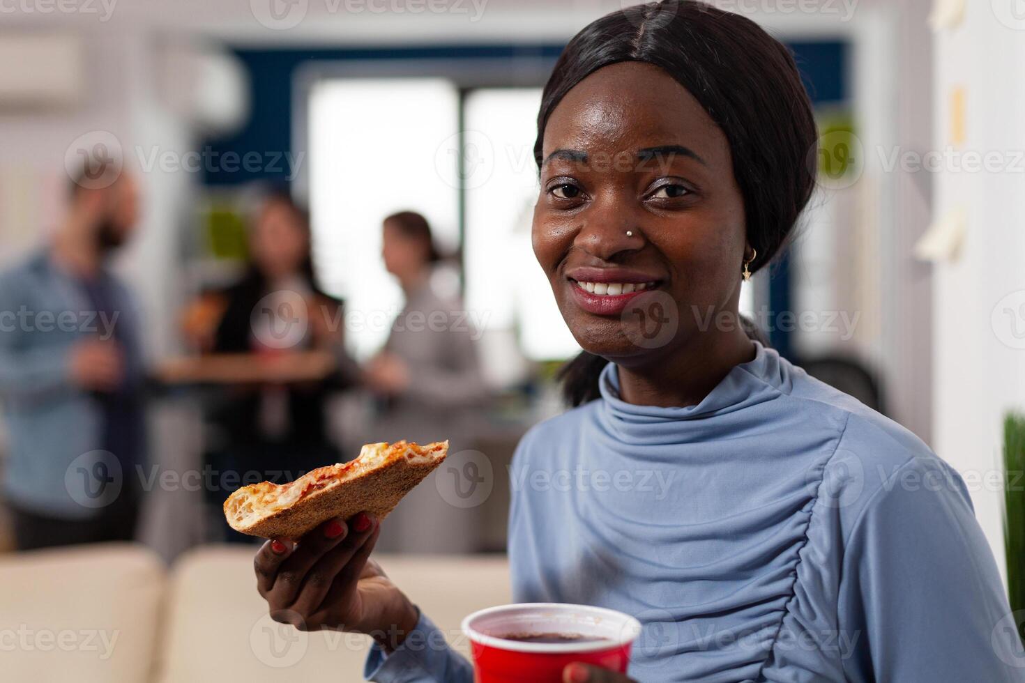 Portrait of african american woman holding pizza and beer, enjoying after work drinks party with office coworkers. Cheerful person having fun at celebration after hours with alcohol and snacks. photo