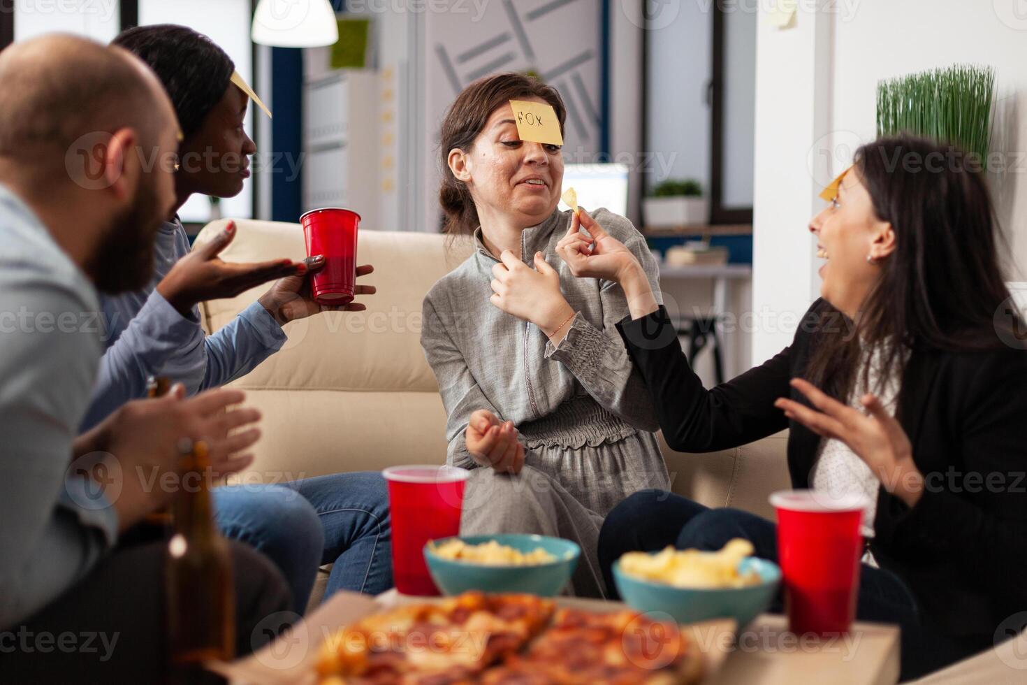 Diverse group of coworkers playing guess who game with sticky notes on forehead, enjoying fun activity with charades play. People guessing pantomime at drinks celebration after work hours. photo