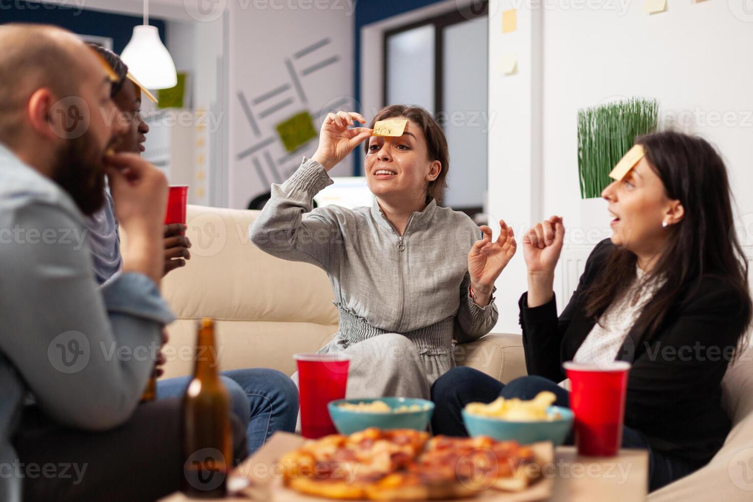 Businesspeople playing charades game and having fun together at office party, using sticky notes on forehead for guessing activity. Man and women enjoying drinks after work hours. photo