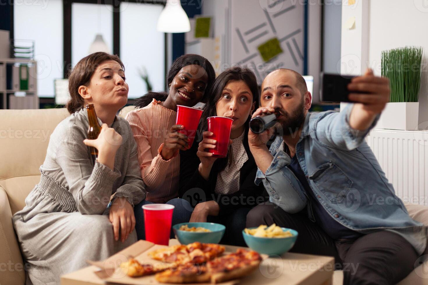 Group of people taking selfies and celebrating with drinks in office after work hours. Man and women having fun with pictures on smartphone, enjoying alcoholic drinks and snacks at party. photo