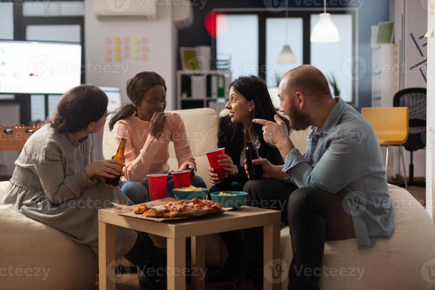Multiethnic group of coworkers having conversation at office celebration. Man and women talking and having fun with alcoholic drinks, pizza and chips at party after work hours. photo
