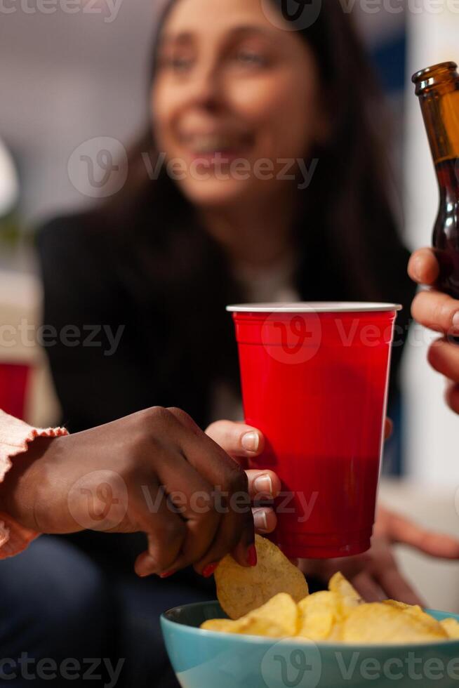 Cheerful people enjoying office party celebration with drinks and snacks after work hours. Coworkers eating chips from bowl, holding glasses and bottles of beer, doing fun activity. Close up. photo