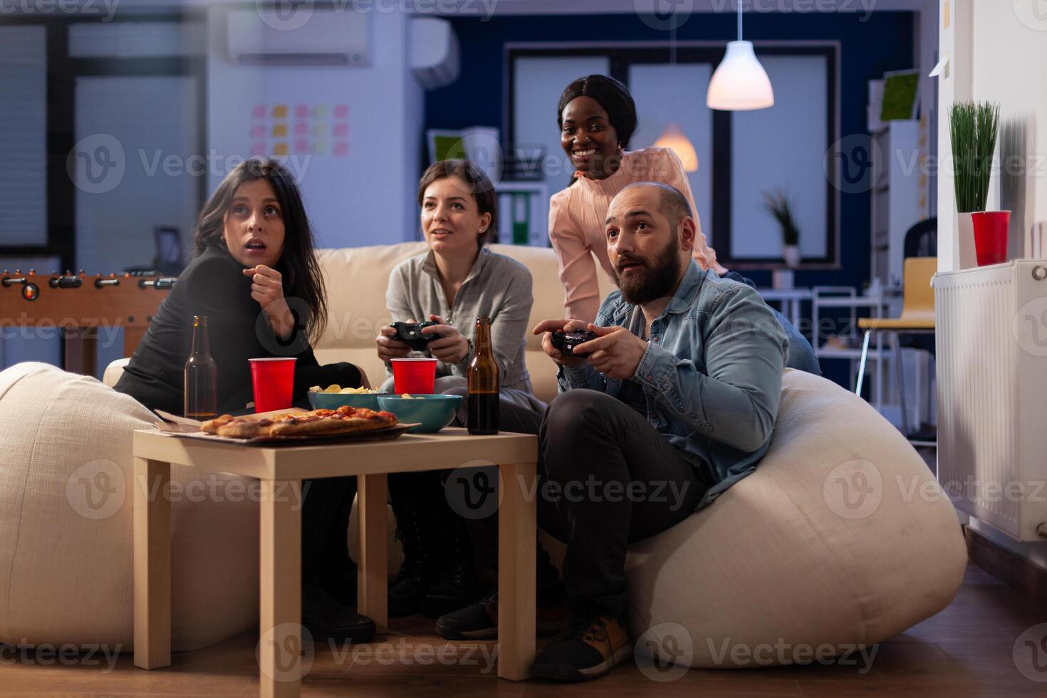 Smiling people playing games online on tv console, using controller to win at office part after work. Colleagues having fun with game, eating pizza and drinking alcoholic beer. photo