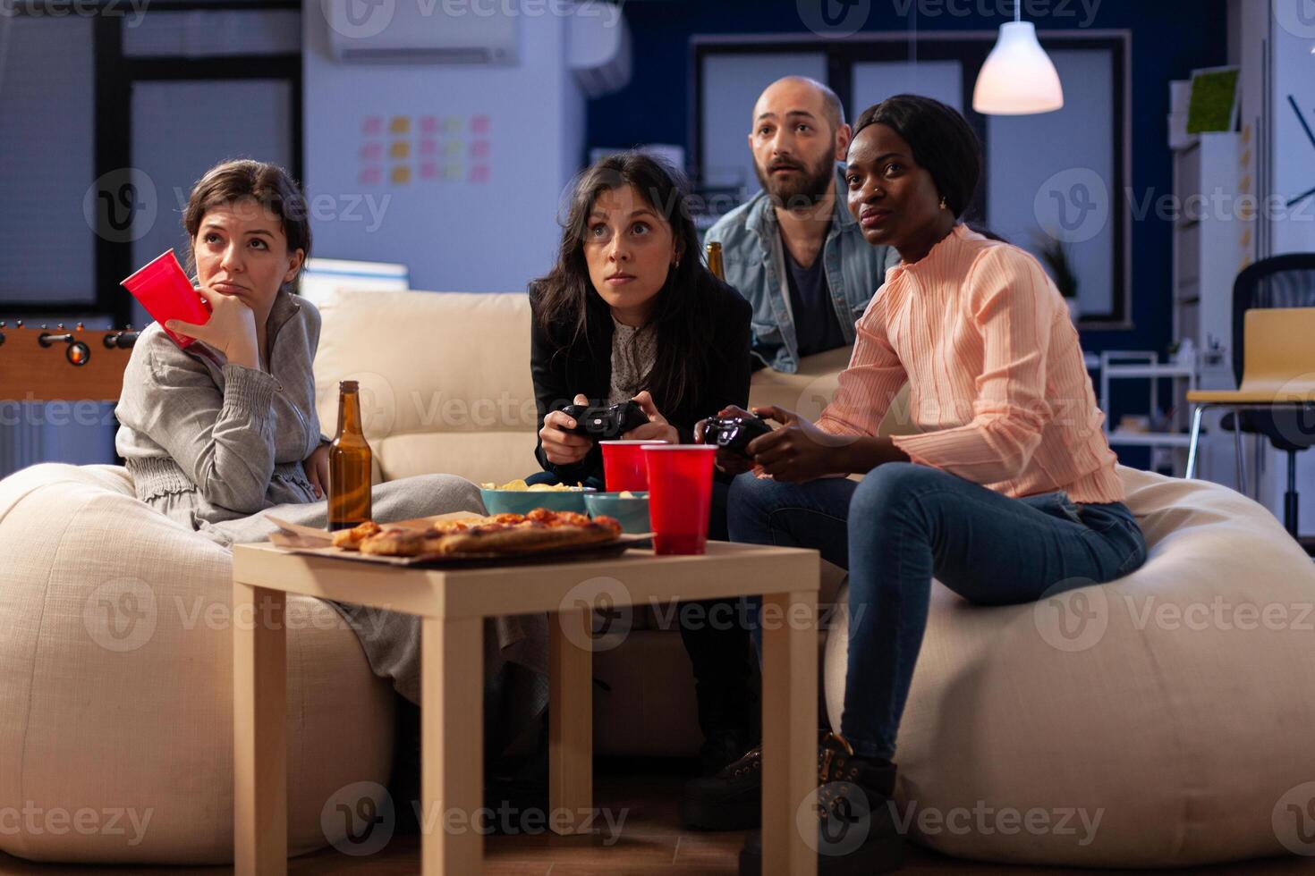 Diverse group of coworkers having fun with games on console, enjoying drinks and snacks at office celebration after work hours. People using controller to play online game at gathering. photo