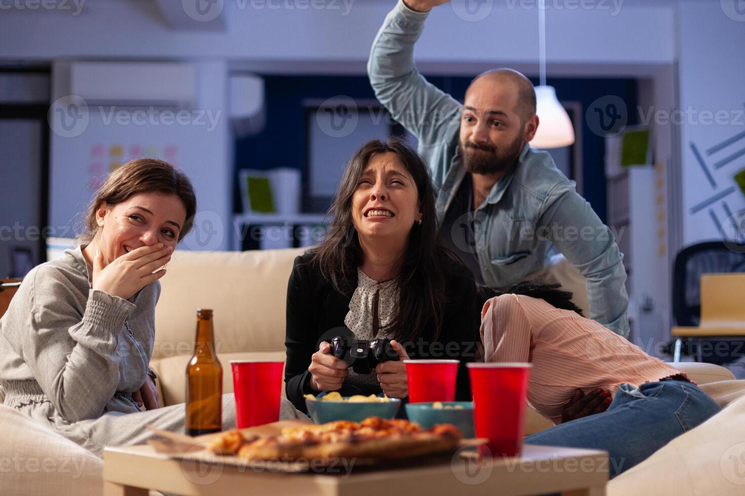 Office coworkers feeling frustrated about lost games with controller on console, doing leisure activity at celebration after work. Business people enjoying party with drinks. photo