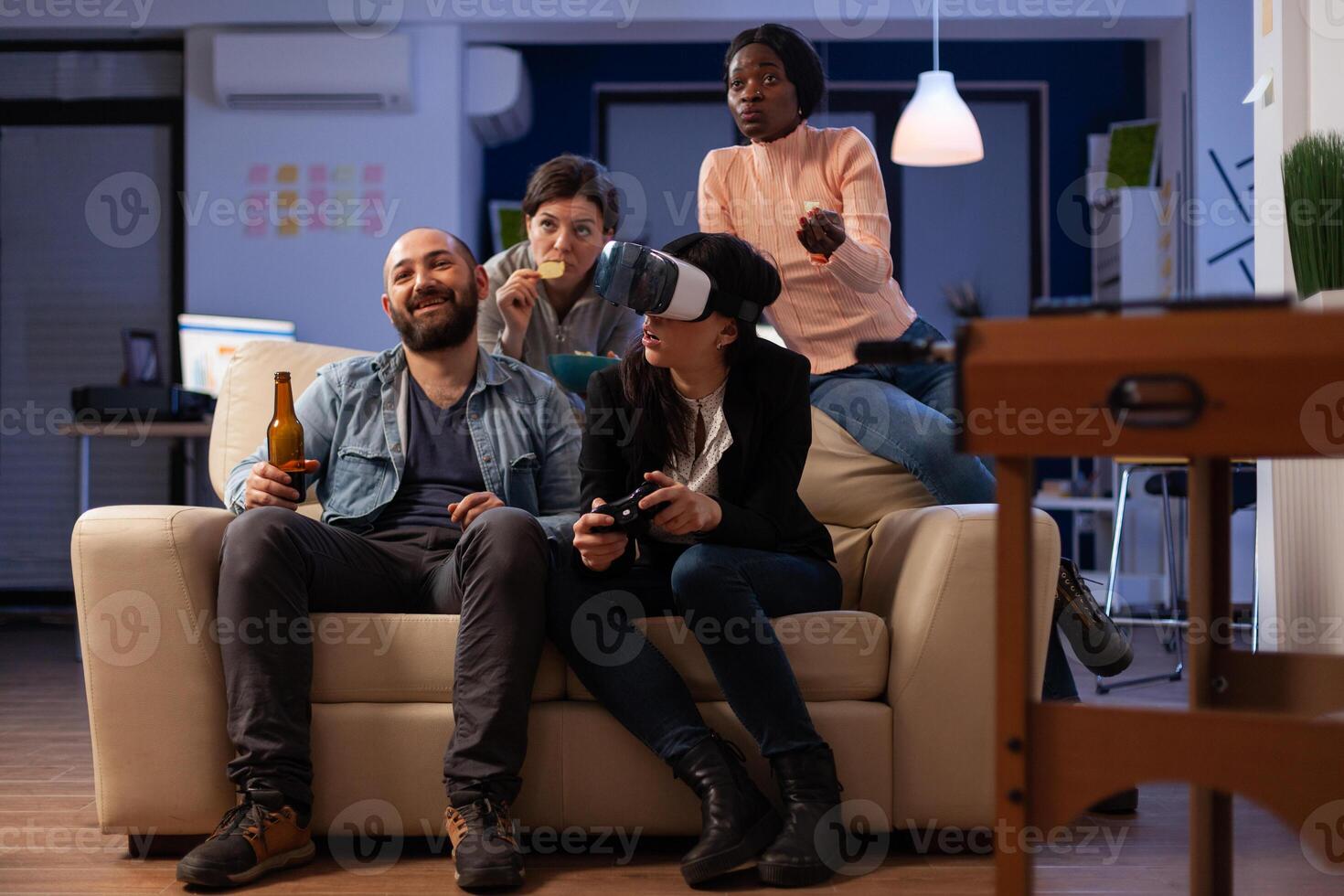 Multiethnic group of people having fun with games on console and virtual reality glasses. Coworkers meeting at office party celebration with beer drinks after work hours. photo