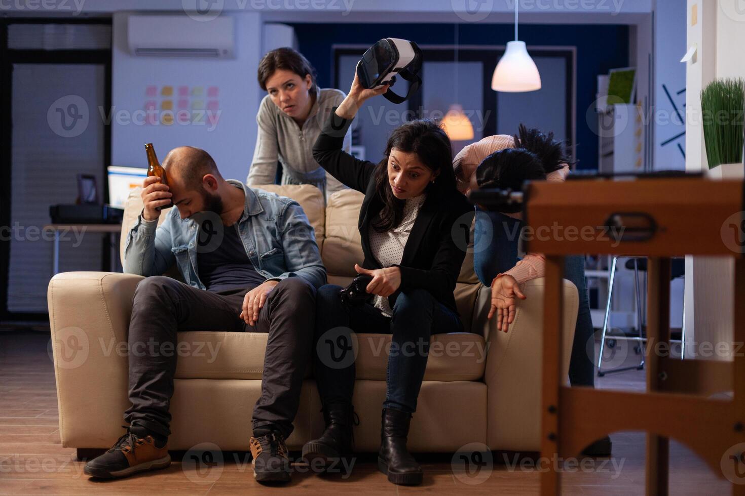 Diverse group of people feeling sad about lost games with virtual reality glasses on console. Man and women meeting for drinks after work, doing leisure activity in business office. photo