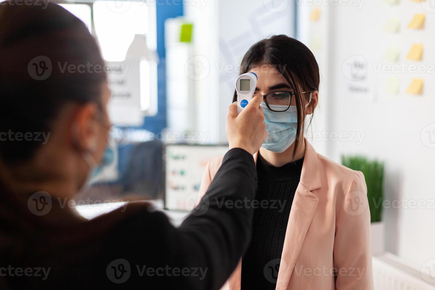 Woman checking colleague temperature using digital thermometer in work office during global pandemic with coronavirus, wearing face mask as safety precaution. New normal virus outbreak. photo