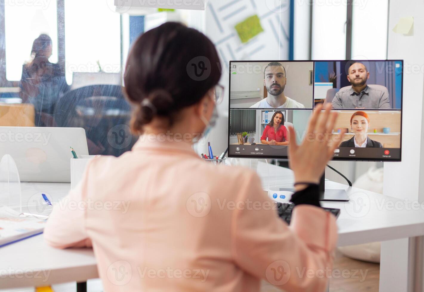 Executive woman in new nomal workplace talking on remote call with team, wearing face mask due to coronavirus flu outbreak. Looking at monitor screen during virtual call. photo