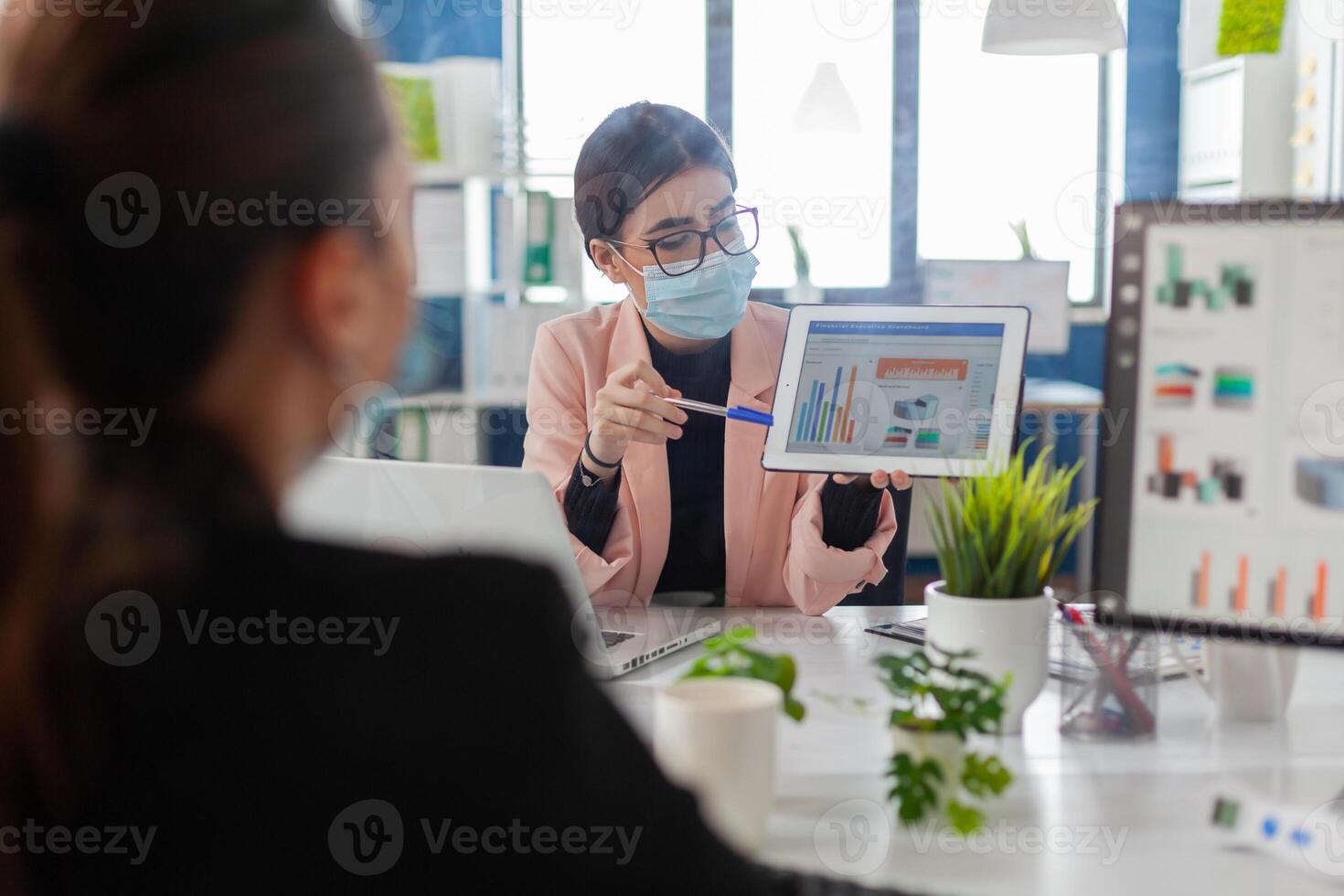Close up of coworkers with face mask working together at financial project using tablet computer while sitting in company office. Team mantain social distance to avoid infection with covid19. photo