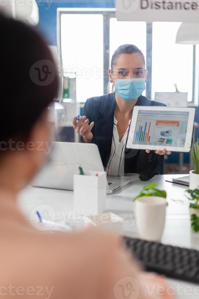 Executive manager with face mask showing statistics to her collegue using digital tablet computer. Coworkers working in new normal office keeping social distance to prevent infection with covid19. photo