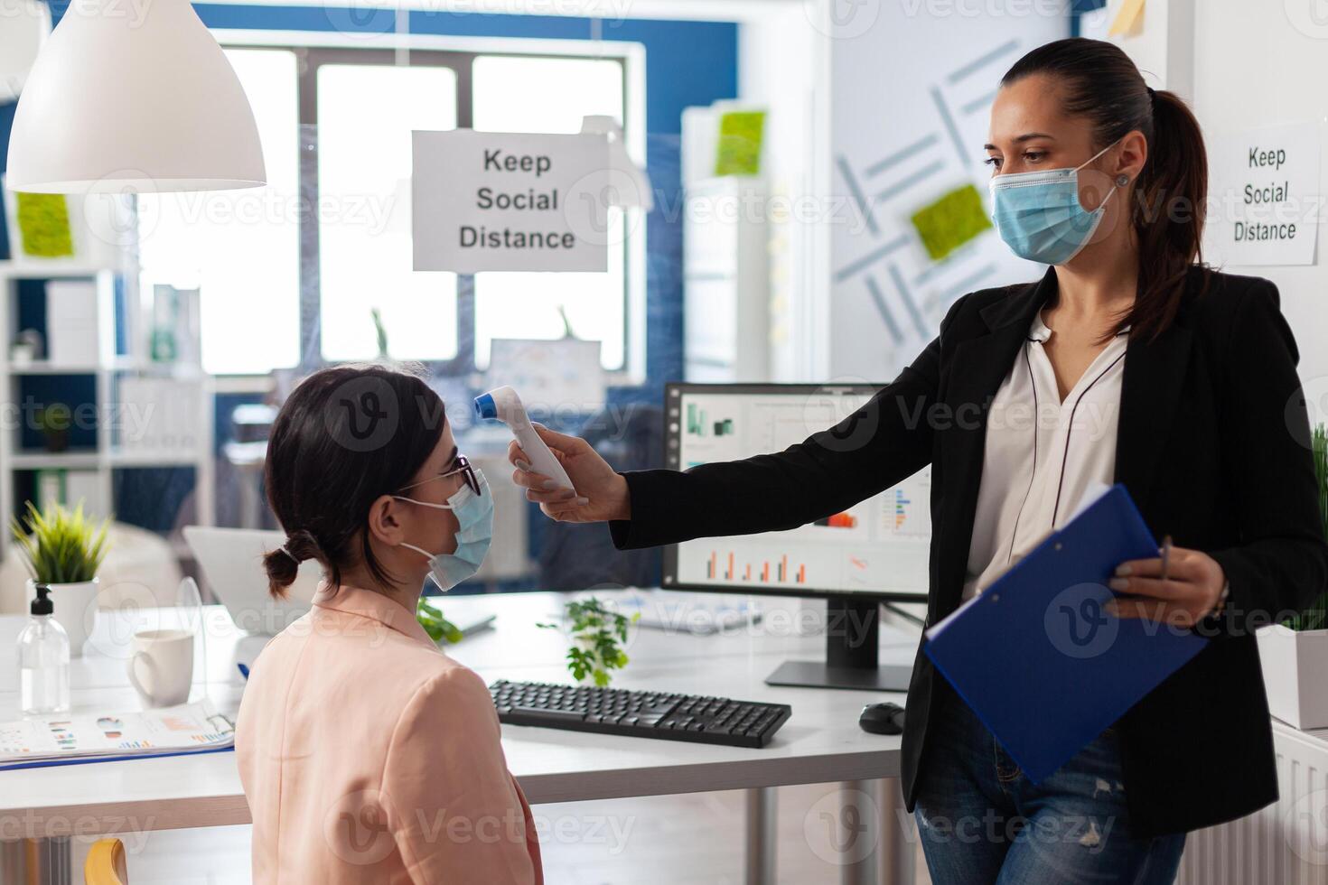 Manager using medical thermometer checking collegue temperature to prevent infection with coronavirus during global pandemic in startup business office. Teamwork with face mask against covid19 photo