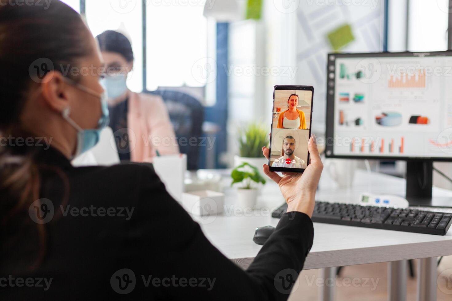 Businesswoman holding smartphone discussing with remote friends having online videocall conference meeting working in company office. Manager with face mask against covid19 during teleconference photo
